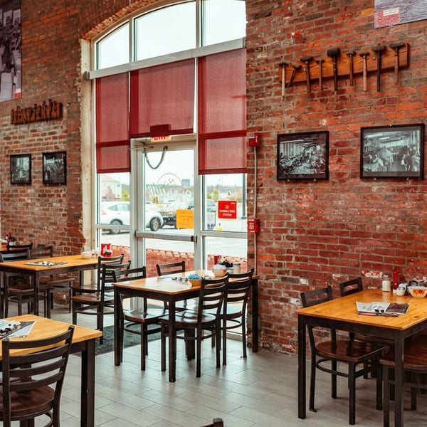 Restaurant interior with brick walls, wooden tables, chairs, and large windows with red blinds.