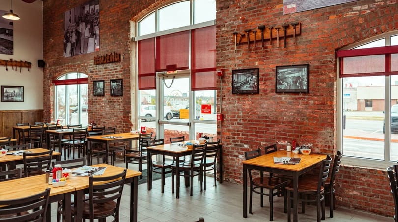 Restaurant interior with brick walls, wooden tables, chairs, and large windows with red blinds.