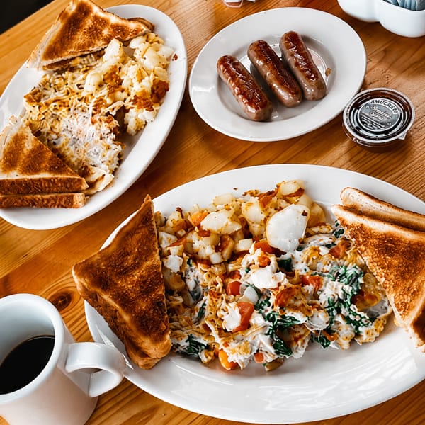 Breakfast spread with scrambled eggs, home fries, toast, sausages, and coffee on a wooden table.