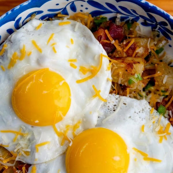 Two fried eggs with bright yellow yolks on a breakfast bowl with potatoes, bacon, and cheese.