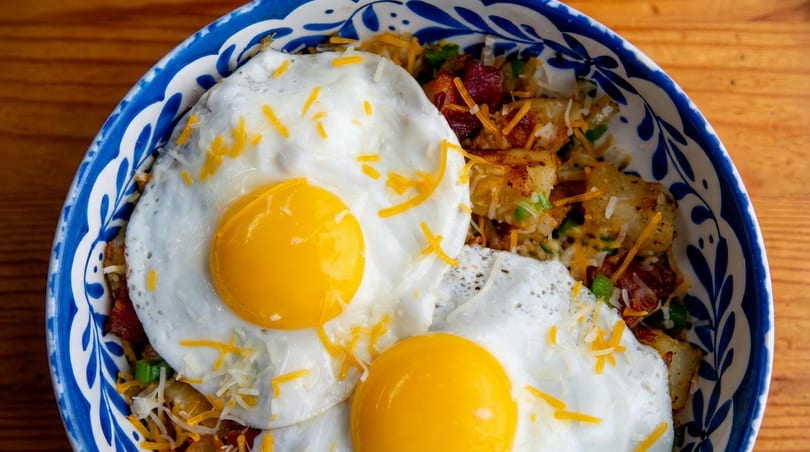 Close-up of a breakfast bowl with fried eggs, potatoes, bacon, and cheese in a blue and white bowl.
