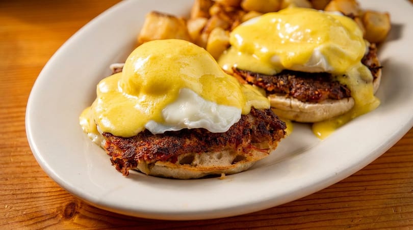 Two crab cake eggs Benedict with hollandaise sauce and a side of roasted potatoes on a white plate.