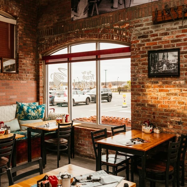 Cozy restaurant interior with brick walls, wooden tables, cushioned bench seating, and large windows overlooking a parking lot.