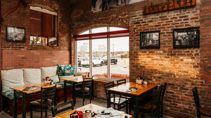 Cozy restaurant interior with brick walls, wooden tables, cushioned bench seating, and large windows overlooking a parking lot.