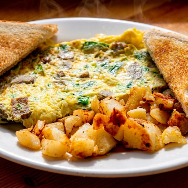 Steaming omelet with meat and greens, home fries, and toast on a white plate.