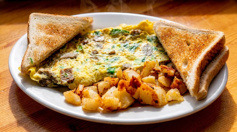 Steaming omelet with meat and greens, home fries, and toast on a white plate.