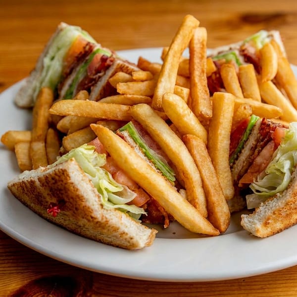 A white plate holds a club sandwich cut into quarters, surrounded by golden french fries on a wooden table.