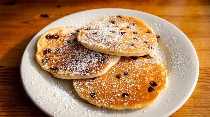Three golden-brown chocolate chip pancakes dusted with powdered sugar on a white plate.