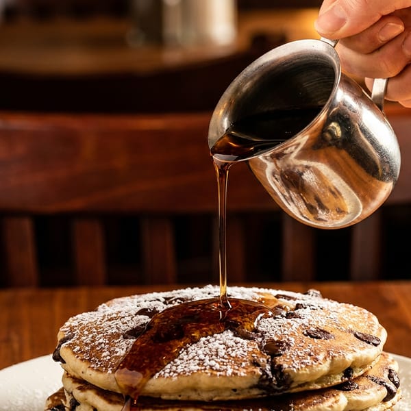 A hand pours maple syrup from a silver pitcher onto a stack of chocolate chip pancakes.