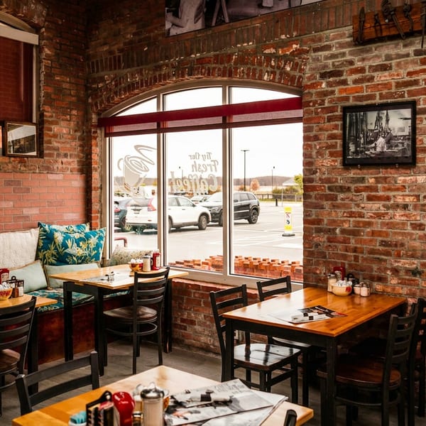 Cozy restaurant interior with brick walls, wooden tables, booth seating, and large windows overlooking a parking lot.