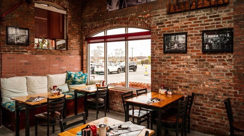 Cozy restaurant interior with brick walls, wooden tables, booth seating, and large windows overlooking a parking lot.